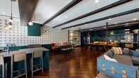Located at 311 El Camino Real in San Carlos, California, this thoughtfully designed single-floor amenity space blends industrial character with modern comfort. Exposed dark wooden ceiling beams run throughout, complementing the striking herringbone hardwood flooring that anchors the open-plan layout. Black steel shelving units divide the space into distinct functional zones while maintaining an airy, connected feel.

The kitchen area is a standout feature, equipped with a professional Wolf gas range cooktop, bold teal cabinetry, dark stone countertops, and a decorative patterned tile backsplash that adds visual interest. Adjacent dining areas offer communal long tables and chairs, ideal for gatherings. A spacious lounge zone features sectional seating, including a distinctive blue sofa with trunk-style detailing, alongside casual bean bag seating for a relaxed atmosphere.

A cozy sitting area near large black-framed French doors opens to views of an outdoor pool courtyard, flooding the space with natural light. A separate lounge nook features a herringbone wood-paneled fireplace wall, a wall-mounted display screen, framed artwork, and plush accent seating. Throughout, rich teal accent walls, patterned wallcoverings, and lush indoor plants contribute to a vibrant yet refined communal environment.