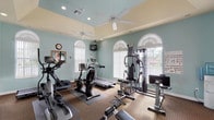 This fitness center is thoughtfully designed to support a full range of workout activities within a single, open floor plan. The space is painted in soft sage green and cream tones, creating a calm and inviting atmosphere conducive to exercise. Arched windows with fan-style transoms line the walls, allowing ample natural light to fill the room while offering views of the surrounding outdoor greenery. Ceiling fans are mounted above to maintain comfortable airflow throughout workouts.

The cardio area is equipped with treadmills, an elliptical trainer, and a stationary recumbent bike, providing multiple options for aerobic exercise. A multi-station cable machine anchors the strength-training section, complemented by a free weight area featuring dumbbells, a flat bench, and an adjustable weight bench. A padded exercise mat and weight step platform offer space for floor-based training.

Floor-to-ceiling mirrors span one full wall, visually expanding the space and allowing users to monitor their form. Recessed lighting supplements the natural light. A wall-mounted television provides entertainment during workouts, and a water cooler station is conveniently positioned near the entrance. The carpeted flooring with rubber equipment mats ensures stability and comfort throughout the space.