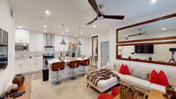 This thoughtfully designed single-floor residence offers a seamless open-concept layout that connects the kitchen, dining, and living areas into one cohesive and inviting space. The kitchen is a standout feature, showcasing crisp white shaker cabinetry, striking marble-veined quartz countertops, a large center island with bar seating, stainless steel appliances including a range with a sleek column hood, and industrial-style pendant lighting. Light wood-tone flooring runs throughout the main living area, complementing the bright, recessed-lit ceiling equipped with a modern ceiling fan. The living room offers a comfortable, open atmosphere with warm accent décor and large mirrors that enhance the sense of space. Each bedroom is generously sized with plush carpeting, ceiling fans, natural light from slatted blinds, and ample closet space. The primary bedroom includes a walk-through closet leading to a well-appointed en suite bathroom. Both bathrooms feature elegant marble-topped vanities with under-mount sinks and LED-backlit mirrors, adding a clean, contemporary touch. A glass-enclosed shower is visible in the primary bath. The overall design balances modern finishes with warm, mid-century inspired furnishings, creating a polished and functional living environment.