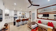 This thoughtfully designed single-floor residence offers a seamless open-concept layout that connects the kitchen, dining, and living areas into one cohesive and inviting space. The kitchen is a standout feature, showcasing crisp white shaker cabinetry, striking marble-veined quartz countertops, a large center island with bar seating, stainless steel appliances including a range with a sleek column hood, and industrial-style pendant lighting. Light wood-tone flooring runs throughout the main living area, complementing the bright, recessed-lit ceiling equipped with a modern ceiling fan. The living room offers a comfortable, open atmosphere with warm accent décor and large mirrors that enhance the sense of space. Each bedroom is generously sized with plush carpeting, ceiling fans, natural light from slatted blinds, and ample closet space. The primary bedroom includes a walk-through closet leading to a well-appointed en suite bathroom. Both bathrooms feature elegant marble-topped vanities with under-mount sinks and LED-backlit mirrors, adding a clean, contemporary touch. A glass-enclosed shower is visible in the primary bath. The overall design balances modern finishes with warm, mid-century inspired furnishings, creating a polished and functional living environment.