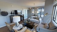 This single-floor space showcases a well-appointed living area designed with a contemporary aesthetic. The room is anchored by a large sectional sofa in crisp white upholstery, complemented by patterned accent pillows in grey and blue tones, alongside a houndstooth-upholstered armchair and a tan leather seating piece. A dark espresso square coffee table sits atop a plush shag area rug, providing a striking contrast against the light hardwood flooring that runs throughout.

Floor-to-ceiling windows along one wall flood the space with abundant natural light while offering views of the surrounding urban environment. The windows also feature access to a balcony or outdoor area, enhancing the open, airy feel of the room.

A built-in media unit houses a large flat-screen television, with open shelving providing functional storage. Overhead, an industrial-style exposed track lighting fixture and a pendant chandelier with globe bulbs add layered illumination. A frosted sliding glass door offers a stylish partition to an adjacent space. Decorative table lamps with chrome bases and white shades contribute to the warm, inviting ambiance of this thoughtfully designed interior.