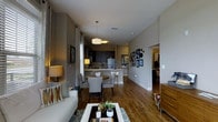 This well-appointed single-floor residence offers a thoughtfully designed open-concept layout that effortlessly connects the main living spaces. Rich hardwood flooring flows throughout, anchoring the warm and inviting atmosphere of the home.

The living area is comfortably furnished and bathed in natural light from large windows dressed with elegant curtains. Decorative accents, including framed artwork and accent pillows, add personality and character to the space.

The dining area sits at the heart of the home, featuring a round table with upholstered chairs positioned beneath a pair of modern pendant lights, creating a cozy and functional gathering spot.

The kitchen is finished with dark cabinetry, a classic tile backsplash, stainless steel appliances, and a white breakfast bar that offers additional seating and prep space. The contrast between the dark cabinetry and light countertops gives the kitchen a contemporary, polished look.

A gallery wall of framed artwork and decorative pieces adds a curated touch to the transition between the main living space and the hallway, which leads to additional rooms beyond. The neutral gray wall tones throughout the home create a cohesive, modern palette that complements the warm wood flooring and furnishings beautifully.