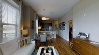 This well-appointed single-floor residence offers a thoughtfully designed open-concept layout that effortlessly connects the main living spaces. Rich hardwood flooring flows throughout, anchoring the warm and inviting atmosphere of the home.

The living area is comfortably furnished and bathed in natural light from large windows dressed with elegant curtains. Decorative accents, including framed artwork and accent pillows, add personality and character to the space.

The dining area sits at the heart of the home, featuring a round table with upholstered chairs positioned beneath a pair of modern pendant lights, creating a cozy and functional gathering spot.

The kitchen is finished with dark cabinetry, a classic tile backsplash, stainless steel appliances, and a white breakfast bar that offers additional seating and prep space. The contrast between the dark cabinetry and light countertops gives the kitchen a contemporary, polished look.

A gallery wall of framed artwork and decorative pieces adds a curated touch to the transition between the main living space and the hallway, which leads to additional rooms beyond. The neutral gray wall tones throughout the home create a cohesive, modern palette that complements the warm wood flooring and furnishings beautifully.