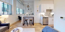 This single-floor residence showcases an open-concept layout that thoughtfully integrates the living, dining, and kitchen areas into one cohesive space. Industrial loft-inspired design elements are prominent throughout, with exposed HVAC ductwork running along the ceiling contrasting elegantly against clean white walls and warm hardwood flooring.

The living area is anchored by a bank of large, multi-pane windows that flood the space with natural light and offer views of the surrounding urban streetscape. A dark leather sofa, low-profile media console, and a contemporary pendant light contribute to the modern aesthetic.

The kitchen is well-appointed with white shaker-style cabinetry, granite countertops with a complementary backsplash, and a full suite of stainless steel appliances including a gas range, over-the-range microwave, French door refrigerator, and dishwasher. A raised breakfast bar with bar stools provides a casual dining space that bridges the kitchen and living zones.

The bedroom, visible in the floor plan, is a distinct private retreat separated from the main living area. The bathroom features large-format marble-look tile flooring, a soaking tub, and a clean, minimalist design with crisp white finishes throughout.