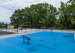 Located at 4570 St Marys Rd in Columbus, Georgia, this outdoor recreational space serves as a central amenity area within a residential community. The grounds are well-maintained and thoughtfully designed to accommodate a variety of outdoor activities for residents of all ages.

The playground area is a standout feature, offering a multi-level wooden play structure complete with a bold blue spiral slide, yellow slide, climbing elements, and colorful panels. Swing sets are positioned nearby, and the entire play zone is surfaced with wood chip mulch for safety and maintenance. Smaller ride-on play features are also scattered across the grassy areas, catering to younger children.

Adjacent to the playground, a swimming pool is enclosed within a chain-link fence lined with green privacy screening. The pool area is outfitted with lounge chairs and shaded by umbrellas, creating a relaxed environment for recreation and leisure. The pool deck provides ample space for sunbathing and socializing.

The surrounding landscape is lush with tall mature trees, including prominent pine trees that provide natural shade and a sense of seclusion. Open green lawns connect the various activity zones, giving the space an expansive, park-like atmosphere. The residential buildings of the community are visible in the background, reinforcing the convenience and accessibility of these amenities for residents.