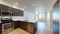 This single-floor space showcases a contemporary design aesthetic with a clean, minimal palette carried consistently across every room. The open-concept layout connects the kitchen and living area seamlessly, creating a generous, light-filled environment anchored by light hardwood flooring that runs throughout the main living spaces.

The kitchen is a standout feature, equipped with flat-front dark cabinetry that extends to the upper walls for substantial storage capacity. A large peninsula with a crisp white quartz countertop provides both prep space and a natural division between the kitchen and living area. Stainless steel appliances — including a refrigerator, gas range, microwave, and dishwasher — complement the dark cabinetry. Recessed lighting illuminates the space, while a large window at the far end of the living area draws in natural light and frames an outdoor view.

The bathroom maintains the same modern sensibility, featuring a dark-toned vanity with a white countertop, an undermount sink, a backlit mirror, and large-format light tile flooring. The bedroom is a well-proportioned, quiet space with the same hardwood flooring and a sliding-door closet offering practical built-in storage. In-unit laundry is also present, adding a notable convenience to this thoughtfully designed space.