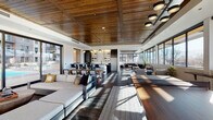 This single-floor clubhouse is a thoughtfully designed communal space that blends contemporary aesthetics with warm, natural materials. The interior is anchored by a striking wood-paneled ceiling, which adds texture and warmth throughout the open-concept layout. Dark hardwood-style flooring runs underfoot, providing a rich contrast to the bright white walls and abundant natural light.

The space is organized into several distinct yet connected zones. A generous lounge area features plush sectional sofas and modern accent chairs arranged around area rugs, creating an inviting atmosphere for relaxation or socializing. A separate seating nook along a full bank of floor-to-ceiling windows frames sweeping mountain and cityscape views, ideal for quiet conversation or enjoying natural light. A tufted leather sofa anchors another lounge corner bathed in sunlight.

The dining area accommodates a large wood table with modern chairs, suitable for group gatherings. Adjacent to it, a well-appointed wet bar features a dramatic marble-veined quartz backsplash, matte black cabinetry with brass hardware, an undermount sink, and open glass shelving with copper pipe accents—adding a refined, industrial touch.

Large sliding glass doors open directly to an outdoor terrace, further extending the usable space and enhancing the connection to the surrounding environment.