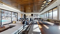 This single-floor clubhouse is a thoughtfully designed communal space that blends contemporary aesthetics with warm, natural materials. The interior is anchored by a striking wood-paneled ceiling, which adds texture and warmth throughout the open-concept layout. Dark hardwood-style flooring runs underfoot, providing a rich contrast to the bright white walls and abundant natural light.

The space is organized into several distinct yet connected zones. A generous lounge area features plush sectional sofas and modern accent chairs arranged around area rugs, creating an inviting atmosphere for relaxation or socializing. A separate seating nook along a full bank of floor-to-ceiling windows frames sweeping mountain and cityscape views, ideal for quiet conversation or enjoying natural light. A tufted leather sofa anchors another lounge corner bathed in sunlight.

The dining area accommodates a large wood table with modern chairs, suitable for group gatherings. Adjacent to it, a well-appointed wet bar features a dramatic marble-veined quartz backsplash, matte black cabinetry with brass hardware, an undermount sink, and open glass shelving with copper pipe accents—adding a refined, industrial touch.

Large sliding glass doors open directly to an outdoor terrace, further extending the usable space and enhancing the connection to the surrounding environment.