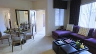 This single-floor residential space offers a warm and inviting layout designed for comfortable everyday living. The living area serves as the heart of the home, featuring plush wall-to-wall carpeting that adds a sense of warmth underfoot. A bold purple sectional sofa, accented with decorative throw pillows, anchors the room alongside a stylish dark ottoman coffee table, creating a striking contemporary aesthetic. Vertical blinds and ornate dark valances frame the windows, allowing natural light to filter in while adding a dramatic design touch to the space.

Adjacent to the living area, a compact dining space accommodates a small table and chairs, conveniently positioned near a large mirror that enhances the sense of openness. The neutral, light-toned walls throughout provide a clean backdrop that complements the rich, jewel-toned furnishings.

A standout feature of this space is the direct access to a covered outdoor patio through a full-height door, seamlessly connecting the interior to the outside. The patio area, visible through the open doorway, offers an extension of the living space ideal for relaxing outdoors. Overall, the space balances bold decorative choices with functional, well-organized living areas.