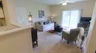 This single-floor residence offers a comfortable and functional open-concept layout designed for everyday living. The living room serves as the heart of the space, featuring plush wall-to-wall carpeting that adds warmth underfoot. A large sliding glass door dominates one wall, flooding the room with abundant natural light and providing a clear view of the outdoor surroundings. The ceiling fan overhead ensures year-round comfort and adds a classic, practical touch to the room's design. The living area is generously proportioned, offering ample room for a full sofa, accent seating, and a central coffee table, all arranged on a colorful area rug that anchors the space. Neutral-toned walls create a versatile backdrop that complements a wide range of décor styles. A partial counter or bar-height divider is visible at the edge of the living area, suggesting a seamless transition into an adjacent kitchen or dining space. This design choice promotes an open flow between the main living areas while maintaining a sense of defined zones. Overall, the space presents a bright, airy, and inviting atmosphere well-suited for both relaxing and entertaining.