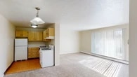 This single-floor residence offers a practical and comfortable layout designed for everyday living. The open-concept living area is carpeted and filled with natural light streaming through large vertical-blind-covered sliding doors, creating a bright and welcoming atmosphere. Baseboard heating runs throughout, ensuring consistent warmth across the space.

The kitchen is efficiently designed with light wood cabinetry offering generous storage, white appliances including a gas range, dishwasher, and refrigerator, complemented by white countertops and parquet-style flooring. A pendant light fixture adds a functional accent overhead.

The bedrooms are carpeted and well-proportioned, each featuring windows with blinds that allow natural light and outdoor views. Wall-unit air conditioners and baseboard heaters provide individual climate control. Closets with sliding doors offer convenient built-in storage.

The bathroom is clean and functional, featuring a tiled tub and shower enclosure with grab bars, a vanity with a wide countertop sink, a mirrored medicine cabinet, and a distinctive mosaic tile floor in neutral multicolor tones. The soft, neutral cream tones used throughout the unit lend a cohesive and understated aesthetic to the entire space.