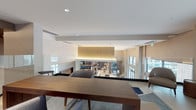 This business center is designed with a contemporary aesthetic that balances functionality and sophisticated style. The interior showcases an open, multi-level layout connected by glass panel railings that allow sightlines across the entire space, fostering an open and collaborative atmosphere. Warm ambient lighting with cove and recessed fixtures highlights the clean-lined architecture, complemented by neutral tones of white, taupe, and deep navy accent walls. The interior features well-appointed seating areas with modern furniture, including wood-tone tables and upholstered chairs, arranged to support both focused work and informal meetings. A presentation area equipped with a projector screen and display stand provides a dedicated zone for group sessions or demonstrations. Large windows allow natural light to filter throughout, enhancing the bright and productive environment. The exterior transitions seamlessly to a covered outdoor terrace supported by natural stone columns and finished with large-format tile flooring. A warm wood-paneled ceiling and ceiling fans overhead create a comfortable, shaded retreat. The terrace opens to views of a pool area and surrounding landscaping, offering a refreshing outdoor setting ideal for breaks or casual conversations. This business center provides a well-rounded, professional space suitable for a variety of work and meeting needs.