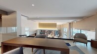 This business center is designed with a contemporary aesthetic that balances functionality and sophisticated style. The interior showcases an open, multi-level layout connected by glass panel railings that allow sightlines across the entire space, fostering an open and collaborative atmosphere. Warm ambient lighting with cove and recessed fixtures highlights the clean-lined architecture, complemented by neutral tones of white, taupe, and deep navy accent walls. The interior features well-appointed seating areas with modern furniture, including wood-tone tables and upholstered chairs, arranged to support both focused work and informal meetings. A presentation area equipped with a projector screen and display stand provides a dedicated zone for group sessions or demonstrations. Large windows allow natural light to filter throughout, enhancing the bright and productive environment. The exterior transitions seamlessly to a covered outdoor terrace supported by natural stone columns and finished with large-format tile flooring. A warm wood-paneled ceiling and ceiling fans overhead create a comfortable, shaded retreat. The terrace opens to views of a pool area and surrounding landscaping, offering a refreshing outdoor setting ideal for breaks or casual conversations. This business center provides a well-rounded, professional space suitable for a variety of work and meeting needs.
