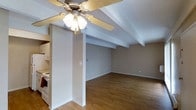 Located at 7100 East Evans Avenue in Denver, Colorado, this single-floor space offers a straightforward and functional layout with a clean, understated aesthetic. The open living area is a standout feature, showcasing warm-toned hardwood flooring that runs throughout and lends a sense of continuity and warmth to the interior. Exposed ceiling beams add a subtle architectural character, complementing the neutral, light gray walls that create a bright and versatile backdrop. A ceiling fan with an integrated light fixture is centrally positioned, providing both comfort and illumination to the main living space. A structural column defines the transition between the kitchen and living areas, contributing to the layout's natural flow while maintaining an open feel. The kitchen, partially visible from the main living area, includes white cabinetry and standard appliances, offering practical functionality. White baseboards and trim throughout provide a crisp contrast to the warm flooring and muted wall tones. The space presents a solid foundation that is well-suited for a variety of uses, with its neutral palette and durable finishes making it both adaptable and easy to maintain.