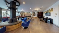 Located at 1493 Osage St in Denver's vibrant 80204 corridor, this dynamic single-floor amenity space is designed to energize and entertain. The open-concept layout features warm wood-tone flooring throughout, with recessed lighting and striking circular pendant fixtures illuminating each zone.

The game and recreation area is a standout highlight, equipped with a pool table, ping-pong table, shuffleboard, and dual wall-mounted flat-screen TVs flanked by sleek gaming consoles. A sculptural circular hanging lounge pod with cushioned seating provides a unique social focal point, complemented by bold blue swivel accent chairs nearby.

A dedicated lounge area offers modular seating with vibrant accent pillows, glass coffee tables, and curated wall art — including a large, vivid portrait painting that commands attention. Expansive floor-to-ceiling windows and glass sliding doors flood the space with natural light and provide views of the surrounding Denver streetscape.

The kitchenette station is finished with a geometric 3D tile backsplash set against a bold red and white color-blocked wall, stainless steel appliances, and clean countertops — ideal for casual entertaining. Throughout the space, colorful murals, geometric floor graphics in yellow and blue, and carefully chosen furnishings reinforce a lively, contemporary aesthetic that reflects the energy of the surrounding Denver community.