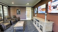 Situated at 5315 Avery Road in Dublin, Ohio, this single-floor space offers a thoughtfully designed interior with a comfortable and polished aesthetic. The standout feature is a dedicated media lounge, finished with deep, warm-toned walls that create an intimate and inviting atmosphere. Crown molding adds a refined architectural detail, while recessed lighting provides a clean, modern overhead illumination throughout the room. A large flat-screen television is wall-mounted above a substantial media console featuring glass-front cabinetry, offering both style and storage. Plush leather reclining seating and a patterned area rug anchor the room with a sense of comfort and sophistication. Classic framed artwork adorns the walls, contributing to the curated feel of the space. French glass doors connect the media room to an adjacent area, allowing natural light to filter through while maintaining a sense of defined spaces. The adjoining room, visible through the doorway, features neutral tones and carpet, offering a contrast to the media lounge's dramatic palette. Overall, the space balances functionality with design, making it well-suited for entertainment and relaxation.