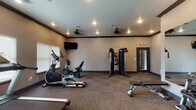 This single-story fitness center offers a comprehensive workout environment designed to accommodate a variety of exercise routines. The space is finished with neutral gray walls, dark crown molding, and durable carpeted flooring throughout, creating a clean and functional aesthetic.

The room is outfitted with a solid selection of equipment, including a treadmill, elliptical trainer, recumbent bike, and a multi-function cable machine for strength training. A three-tier dumbbell rack stocked with a full range of free weights is positioned alongside an adjustable workout bench, supporting both beginner and advanced lifting routines.

A large wall-mounted mirror spans one section of the room, visually expanding the space and allowing users to monitor their form during exercise. Wall-mounted flat-screen televisions are positioned in multiple areas for entertainment during cardio sessions. Ceiling fans and recessed lighting provide comfortable airflow and consistent illumination throughout.

Natural light enters through several windows fitted with horizontal blinds, offering brightness while allowing privacy control. Multiple glass-panel doors provide easy access to and from the space. Overall, the fitness center presents a practical, well-organized environment suited for daily use.