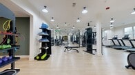 Located at 9821 Research Drive in Charlotte, NC, this fitness center offers a generously sized, single-floor layout designed to accommodate a full range of workout activities. The space features light hardwood-style flooring throughout, white walls accented by deep green painted sections, and a bright, open atmosphere enhanced by large windows that bring in natural light and offer views of the surrounding exterior.

The main workout floor is well-organized and thoughtfully equipped. A dedicated cardio zone houses multiple treadmills, an elliptical, and a stationary rowing machine, all positioned near the windows. A strength training area features multi-station cable machines, adjustable weight benches, and a comprehensive dumbbell rack with accessory storage, including resistance tools like balance domes and foam rollers. Full-length mirrors line key walls, amplifying the sense of space and supporting proper form during workouts.

A separate functional training zone includes a heavy-duty power rack system with battle ropes, resistance bands, and a BOX punching bag, alongside spin bikes and a tablet-enabled smart bike for guided training sessions. Pendant-style lighting fixtures are evenly distributed across the ceiling, ensuring consistent illumination throughout.

A lounge area at one end of the space provides a place to rest, featuring a wall-mounted leather bench, striped seating, and an AED station, rounding out the safety and comfort amenities of this comprehensive facility.