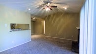 This single-floor residential space offers a straightforward and functional layout designed for comfortable everyday living. The main living area is a notable highlight, featuring wall-to-wall carpeting that adds warmth underfoot and a classic ceiling fan with an integrated light fixture that provides both ambient lighting and air circulation. Recessed track lighting is also present, offering additional illumination options throughout the space.

The living area is open and spacious, with neutral-toned walls that create a calm, versatile backdrop suitable for a variety of interior styles. A pass-through counter connects the living area to the kitchen, promoting an open flow between the two spaces while maintaining a degree of separation. This design element is practical for both entertaining and everyday use.

A hallway visible from the main living area leads to the additional rooms of the home, suggesting a well-organized floor plan. The overall ambiance of the space is clean and understated, with a neutral color palette throughout that makes it easy to personalize. The single-floor layout ensures convenient accessibility, and the combination of functional lighting, open living space, and connected kitchen area makes this a practical and welcoming home.
