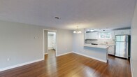 Located at 45 Kings Way in Waltham, Massachusetts, this two-bedroom, one-bathroom home offers a well-organized layout with a thoughtful mix of functional spaces. The combined kitchen and living room serves as the heart of the home, featuring rich hardwood flooring and an elegant chandelier that anchors the living area. The kitchen is outfitted with white cabinetry, light countertops, stainless steel appliances including a refrigerator and dishwasher, and a window that brings in natural light and outdoor views. The open flow between the kitchen and living room creates a comfortable, connected feel throughout the main space. The primary bedroom is generously sized, with hardwood flooring and direct access to the hallway. The second bedroom features plush carpeting and a window that introduces natural light. The full bathroom includes a shower-tub combination with clean, white finishes. A dedicated laundry room with a full-size washer and dryer adds everyday convenience. The staircase, illuminated by a window, connects the levels with clean lines and bright, neutral walls. Throughout the home, soft gray-toned walls, textured ceilings, and crisp white trim contribute to a cohesive and welcoming aesthetic.