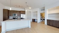 This single-floor residence showcases a thoughtfully designed open-concept layout that seamlessly connects the kitchen and living areas, creating a bright and airy atmosphere. The contemporary kitchen is a standout feature, equipped with stainless steel appliances — including a refrigerator with an ice maker, a built-in microwave, and an electric range — all complemented by rich, dark wood cabinetry with a textured finish. A large central island with a crisp white countertop provides generous workspace and casual seating, while pendant lighting above adds a stylish focal point. A subway tile backsplash adds a subtle layer of texture to the kitchen's clean aesthetic. Light-toned hardwood flooring runs throughout the space, enhancing the sense of openness and warmth. A dedicated built-in desk nook offers a functional workspace tucked neatly between the main living area and the utility room. The in-unit laundry area is conveniently accessible and houses a side-by-side washer and dryer. Recessed and track lighting provide versatile illumination throughout, while white walls and high ceilings contribute to the overall clean, modern ambiance of this well-appointed residence.
