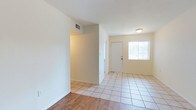 This single-floor space offers a clean, functional layout designed for comfortable everyday living. The entryway greets visitors with large square tile flooring that extends through the main corridor and into the open living areas, providing a bright and easy-to-maintain surface. Adjacent to the entry, warm hardwood-style flooring defines a separate living zone, adding a touch of warmth and contrast to the neutral palette throughout. Crisp white walls and light beige tones create a cohesive, airy ambiance that makes the space feel open and welcoming. A front-facing window near the entryway door allows natural light to filter in, further enhancing the bright atmosphere. The layout includes a defined alcove or hallway that leads to additional rooms, offering a practical separation between the entry and living areas. Ceiling-mounted light fixtures provide consistent illumination throughout. HVAC vents are integrated into the walls, ensuring year-round climate comfort. The overall design is straightforward and well-maintained, making this space a versatile option suitable for a variety of occupants seeking a simple, move-in-ready environment.
