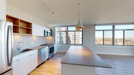 This contemporary single-floor residence showcases a clean, modern design defined by abundant natural light and an open, airy layout. The expansive living area is anchored by floor-to-ceiling windows that wrap around the space, offering sweeping panoramic views of the surrounding cityscape and beyond, while light wood-style flooring adds warmth underfoot.

The open-concept kitchen is thoughtfully designed with sleek white cabinetry, warm wood open shelving, a full suite of stainless steel appliances including a refrigerator, electric range, dishwasher, and microwave, as well as a large central island with neutral countertops. Stylish pendant lighting with gold accents hangs above the island, complementing the track lighting overhead. A tiled backsplash ties the kitchen's contemporary aesthetic together.

The bedrooms are carpeted for comfort, with large windows providing city views and ample closet storage with built-in shelving systems. The hallway connects spaces seamlessly, maintaining the residence's clean, minimalist character.

The bathrooms feature crisp, white finishes, including a soaking tub with subway tile surround and a separate elongated vanity bathroom with large-format tile flooring, an undermount sink, and a full-width mirror. The overall ambiance is bright, modern, and move-in ready.