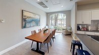 This single-floor space is thoughtfully designed to accommodate a variety of activities within an open, cohesive layout. The lounge area is a standout feature, boasting soaring vaulted ceilings with exposed beam detailing and light-toned hardwood-style flooring throughout. A wall-mounted flat-screen television anchors the seating arrangement of contemporary gray upholstered armchairs gathered around a cylindrical woven side table. Large French doors and expansive windows flood the space with natural light while offering views of the surrounding greenery outdoors.

A dedicated game room area accommodates a full-sized billiards table, providing an additional recreational focal point within the open floor plan. The kitchen is well-equipped with crisp white shaker cabinetry, dark quartz countertops, a subway tile backsplash, and stainless steel appliances, including a large French door refrigerator. Light wood-style flooring continues throughout, maintaining visual continuity.

The dining area features a striking live-edge wood table surrounded by upholstered chairs, accompanied by a large arched window that introduces soft, natural light. A bold abstract painting adorns the wall, adding an artistic element to the refined, neutral-toned interior. Overall, the space exudes a modern yet welcoming ambiance suited for both social gatherings and casual everyday use.