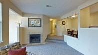 This single-floor residence offers a comfortable and well-connected layout designed for everyday living. The main living area is anchored by a classic tiled fireplace, providing a warm focal point for the room. Soft carpeting underfoot adds a cozy feel throughout the space, complementing the neutral, light-toned walls that create a bright and inviting atmosphere. A few steps lead up to an adjacent hallway, hinting at a thoughtfully split-level flow within the single floor, connecting the living area to additional rooms beyond. The dining area is conveniently positioned just off the living room, separated by a half-wall partition that maintains an open feel while defining each space. A dining table and chairs are naturally accommodated in this area, making it well-suited for daily meals and gatherings. Ceiling-mounted light fixtures provide consistent illumination throughout. A dedicated hallway leads toward what appears to be additional rooms, including utility or storage spaces. Overall, the home presents a practical and comfortable interior with a neutral palette, soft textures, and a layout that promotes easy movement between living, dining, and private areas.