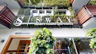 Sway Modern Coastal Apartments presents a distinctive architectural exterior that immediately sets it apart from its urban surroundings. The building's façade features a bold combination of richly toned wood panel cladding arranged in a geometric, mosaic-like pattern, lending warmth and texture to the structure's contemporary design. Contrasting this natural material, a expansive living green wall cascades with dense, verdant foliage across a significant portion of the exterior, reinforcing the coastal and nature-inspired aesthetic the property embodies.

The main entrance is defined by clean white-framed glass doors set beneath a smooth, light-colored stucco finish, creating a welcoming and modern point of arrival. Upper levels feature balconies with metal railings, offering residents outdoor space with views of the surrounding cityscape. The Broadway-facing streetfront includes ground-level glazing that connects the interior to the active pedestrian environment outside.

Located on a well-trafficked urban corridor with neighboring commercial and mixed-use buildings nearby, Sway occupies a prominent position in its neighborhood. The overall design balances organic materials with modern minimalism, creating a cohesive and visually engaging presence that reflects its modern coastal identity.