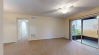 This single-floor unit offers a straightforward, functional layout across several well-defined rooms. The main living area is generously sized, finished with wall-to-wall carpeting in a neutral tan tone that adds warmth underfoot. A ceiling fan with an integrated light fixture sits centrally overhead, complemented by a textured ceiling. Built-in HVAC access panels are mounted along the wall, providing convenient climate control. A sliding glass door along one wall opens to a private balcony, allowing natural light to filter into the room and offering views of surrounding greenery. A hallway connects the living space to the remaining rooms.

The kitchen features rich, medium-toned wood cabinetry extending to the upper walls, providing ample storage. Granite-style countertops run along the perimeter and extend to a peninsula, offering additional prep space. White appliances including a refrigerator, range, and dishwasher are included. Light hardwood flooring and a window above the sink brighten the space.

The bathroom is clean and practical, featuring a wood-toned vanity with a granite-style countertop, a full tub and shower combination with tiled surround, and wood-look flooring. Additional carpeted rooms offer flexible use as bedrooms or flex spaces.