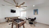 This single-floor game room is designed as a dedicated entertainment space, offering a diverse lineup of recreational activities within a clean, modern interior. The room is finished with light wood-look tile flooring and crisp white walls, creating a bright and open atmosphere complemented by recessed lighting and a large multi-blade ceiling fan that anchors the central area.

The space is well-equipped for group entertainment, featuring a full-size foosball table, a long shuffleboard table, and a classic arcade cocktail cabinet with dual joystick controls and bar-height stools. A dartboard is mounted on one wall alongside an Arizona Wildcats banner and a playful llama canvas print, adding personality and a casual, spirited character to the room.

A wall-mounted flat-screen TV provides additional entertainment options, while a round bistro-style table with modern black chairs offers a comfortable seating area for socializing. Natural light filters in through roller-shaded windows, and a set of French doors with glass panels connects the room to an adjacent exterior area, enhancing the sense of openness. The combination of games, seating, and decor makes this space an ideal venue for gatherings and recreational use.