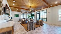 This thoughtfully designed single-floor space encompasses a welcoming entrance, a dining room, and a private office, unified by a cohesive coastal-inspired aesthetic. Throughout the space, striking warm wood-paneled vaulted ceilings with exposed beams draw the eye upward, while decorative patterned tile flooring adds a refined, textural element underfoot. The main gathering area is anchored by a central seating arrangement atop a natural jute rug, surrounded by textured upholstered chairs and illuminated by a geometric chandelier and woven pendant lights. Large black-framed floor-to-ceiling windows and glass doors flood the interior with abundant natural light and provide views of the surrounding outdoor greenery. Lush tropical plants are strategically placed throughout, enhancing the fresh, resort-like ambiance. The dining area features a dark-topped communal table paired with woven rattan chairs and additional pendant lighting, offering a functional yet stylish setting. A built-in wood cabinetry unit with a sink and a wall-mounted television anchors one side of the main room, while a dedicated office nook provides a focused workspace with a wall-mounted wooden desk, ergonomic seating, a brass sconce, and nature-inspired wall art. The overall atmosphere is warm, airy, and refined.