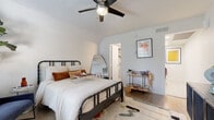 This single-floor space offers a thoughtfully designed layout that seamlessly connects a comfortable bedroom, a compact open-plan living and kitchen area, and a clean, well-appointed bathroom. The bedroom is a standout feature, anchored by a classic black spindle bed frame and dressed in soft, neutral linens. A ceiling fan with an integrated light fixture keeps the room comfortable, while large windows dressed in sheer white curtains flood the space with natural light. A sitting area with deep navy blue accent chairs and a tall fiddle-leaf fig tree adds warmth and character. The bedroom also provides direct access to a walk-in closet area for added storage and convenience. The open-plan living and kitchen zone is bright and functional, with white cabinetry and a layout that maximizes the available footprint. A stylish utility cart and curated wall art contribute to the space's modern bohemian aesthetic throughout. The bathroom is finished with large-format light tile flooring, a clean white vanity, and decorative accents that maintain the cohesive, contemporary feel of the home. Connecting hallways are crisp and well-lit, with recessed lighting and tasteful artwork enhancing the flow between areas. Overall, the space presents a harmonious blend of function and style.