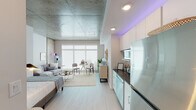 This thoughtfully designed single-floor unit blends industrial and contemporary aesthetics to create a bright, open living environment. The most striking architectural feature is the exposed concrete ceiling, which spans the entire space and lends a distinctive urban character that contrasts beautifully with the warm white walls and light wood-look flooring throughout.

The open-plan living area is anchored by expansive floor-to-ceiling windows that flood the space with natural light, creating an airy and inviting atmosphere. The layout seamlessly accommodates a sleeping area, a comfortable lounge with a sofa and coffee table, and a compact dining nook with a round table and seating — all flowing naturally within a single cohesive space.

The kitchen is efficiently arranged along one wall and features white cabinetry, dark granite countertops, stainless steel appliances including a full-size refrigerator, dishwasher, and built-in microwave, complemented by accent lighting that adds a modern touch. Adjacent to the kitchen, a stacked in-unit washer and dryer offers added convenience.

The bathroom is clean and well-appointed, featuring a tiled walk-in shower with a glass door, a vanity with granite countertops, and light, neutral finishes that maintain the unit's cohesive modern palette. Overall, the space offers a well-balanced combination of functional design and contemporary urban style.