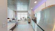 This thoughtfully designed single-floor unit blends industrial and contemporary aesthetics to create a bright, open living environment. The most striking architectural feature is the exposed concrete ceiling, which spans the entire space and lends a distinctive urban character that contrasts beautifully with the warm white walls and light wood-look flooring throughout.

The open-plan living area is anchored by expansive floor-to-ceiling windows that flood the space with natural light, creating an airy and inviting atmosphere. The layout seamlessly accommodates a sleeping area, a comfortable lounge with a sofa and coffee table, and a compact dining nook with a round table and seating — all flowing naturally within a single cohesive space.

The kitchen is efficiently arranged along one wall and features white cabinetry, dark granite countertops, stainless steel appliances including a full-size refrigerator, dishwasher, and built-in microwave, complemented by accent lighting that adds a modern touch. Adjacent to the kitchen, a stacked in-unit washer and dryer offers added convenience.

The bathroom is clean and well-appointed, featuring a tiled walk-in shower with a glass door, a vanity with granite countertops, and light, neutral finishes that maintain the unit's cohesive modern palette. Overall, the space offers a well-balanced combination of functional design and contemporary urban style.