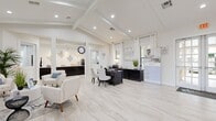 This single-floor space serves as both a welcoming leasing office and a model living showcase, thoughtfully designed to impress visitors from the moment they arrive. The grand entry features soaring ceilings, sleek marble-look porcelain tile flooring, and glass-panel double doors that flood the foyer with natural light. A mounted display near the entrance highlights community amenities, setting an inviting first impression.

The main area is divided into functional zones that flow seamlessly into one another. A professional consultation area features a dark-stained desk, upholstered guest chairs, and storage credenzas, creating an elegant yet functional workspace. Adjacent to this is a comfortable lounge seating area with tufted white armchairs, a round side table, and vibrant blue accent art — ideal for welcoming prospective residents.

Further into the space, an expansive living room showcase boasts exposed beam ceilings, a floor-to-ceiling paneled fireplace wall with a wall-mounted television, and generous seating in neutral tones accented with sky-blue textiles. Multiple windows dressed in white curtains fill the room with natural light.

A fully equipped kitchen features bold dark cabinetry contrasted by bright white quartz countertops, stainless steel appliances, and a center island, demonstrating the quality finishes available throughout the community.