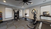 Located at 7 Southpointe Dr in Greenville, South Carolina, this fitness center offers a well-equipped, single-floor workout facility designed to support a wide range of exercise routines. The space is finished with durable patterned carpet tiles in neutral tones of gray and blue, complementing the light gray walls and recessed lighting throughout.

The main workout area is thoughtfully organized to maximize usable floor space. A full rack of dumbbells lines one wall, accompanied by a large wall-mounted mirror that enhances the sense of openness. Cardio equipment includes a treadmill, stationary bike, elliptical, and a stair-stepper, all positioned to take advantage of natural light streaming through multiple windows fitted with horizontal blinds. A multi-station cable and weight machine provides options for strength training, while a rack of medicine balls and a wall-mounted exercise mat offer versatility for functional fitness routines.

Overhead, a large ceiling fan with integrated lighting promotes air circulation during workouts. Contemporary wall art adds a finishing touch to the space. Multiple entry and exit points, including a glass-panel exterior door, ensure easy access. The overall ambiance is clean, functional, and well-maintained, making this fitness center a practical and inviting resource for daily exercise.