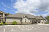 This residential community is designed with a practical, single-story layout spread across a spacious campus-style setting. The exterior architecture features neutral-toned siding, stone accents, and pitched rooflines that lend a cohesive, suburban aesthetic to the development. Multiple buildings are arranged around a network of wide, well-paved driveways that provide easy navigation throughout the property.

Parking is a notable feature of the community, offering both open-air surface parking lots and covered carport structures supported by sturdy columns. The carports provide residents with protected parking options, shielding vehicles from the elements while maintaining an open, airy feel.

The grounds are thoughtfully landscaped, with neatly trimmed shrubs, young deciduous trees, and well-maintained mulched planting beds that add greenery and visual appeal to the paved areas. Manicured lawns extend between the buildings, contributing to the community's clean and welcoming atmosphere.

The open layout of the property allows for ample natural light across all areas, and the surrounding greenery creates a pleasant, park-like environment. The combination of accessible single-floor design, generous parking, and carefully maintained outdoor spaces makes this community a functional and comfortable setting for its residents.