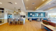 Located on Chief Justice Cushing Highway in Scituate, MA, this single-floor community clubhouse is designed with a coastal New England aesthetic that blends functionality with relaxed, seaside charm. The open-plan layout flows seamlessly from a fully equipped demonstration kitchen to a social dining and lounge area. The kitchen features a large quartz-topped island with bar seating, a stainless steel range hood, built-in wall oven, microwave, full-size refrigerator, and white shaker cabinetry with gold hardware — all complemented by a mosaic tile backsplash and warm pendant lighting. Adjacent to the kitchen, a dining banquette anchors a vibrant teal accent wall adorned with a curated gallery of nautical and coastal artwork. Woven rattan chairs and wood dining tables complete the inviting gathering space. The living area showcases a tufted leather sofa, a linear stone fireplace, a wall-mounted television, and a textured wood-panel tray ceiling, with large windows offering views of the surrounding landscape. A bright sunroom with floor-to-ceiling windows provides direct access to an outdoor patio area. The fitness room is outfitted with cardio equipment, free weights, a stationary bike, and a wall mirror, with natural light from an exterior-facing window. Light hardwood flooring unifies the main areas throughout.