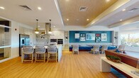 Located on Chief Justice Cushing Highway in Scituate, MA, this single-floor community clubhouse is designed with a coastal New England aesthetic that blends functionality with relaxed, seaside charm. The open-plan layout flows seamlessly from a fully equipped demonstration kitchen to a social dining and lounge area. The kitchen features a large quartz-topped island with bar seating, a stainless steel range hood, built-in wall oven, microwave, full-size refrigerator, and white shaker cabinetry with gold hardware — all complemented by a mosaic tile backsplash and warm pendant lighting. Adjacent to the kitchen, a dining banquette anchors a vibrant teal accent wall adorned with a curated gallery of nautical and coastal artwork. Woven rattan chairs and wood dining tables complete the inviting gathering space. The living area showcases a tufted leather sofa, a linear stone fireplace, a wall-mounted television, and a textured wood-panel tray ceiling, with large windows offering views of the surrounding landscape. A bright sunroom with floor-to-ceiling windows provides direct access to an outdoor patio area. The fitness room is outfitted with cardio equipment, free weights, a stationary bike, and a wall mirror, with natural light from an exterior-facing window. Light hardwood flooring unifies the main areas throughout.