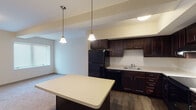 This single-floor unit is thoughtfully designed with a cohesive aesthetic carried throughout each space. The kitchen serves as a focal point, showcasing rich dark espresso cabinetry that extends to the ceiling, offering generous storage. Black appliances, including a refrigerator, range, and dishwasher, pair with light countertops to create a classic contrast. A central island with a matching countertop provides additional workspace and is illuminated by pendant lighting that adds warmth to the area. Wood-style flooring runs throughout the kitchen, transitioning to plush carpet in the adjacent living and bedroom areas.

The living space is open and bright, receiving natural light through windows fitted with blinds. The bedroom features soft carpeting, a tall narrow window, and a spacious walk-in closet equipped with wire shelving and hanging rods, providing ample organization options.

The bathroom is well-finished, featuring a dark vanity with drawers and cabinet storage, a white countertop, a lighted mirror, a medicine cabinet, and a walk-in shower with built-in shelving. Dark hardwood-style flooring completes the bathroom's polished look. The overall palette of warm neutrals and dark wood tones creates a consistent, inviting atmosphere throughout the unit.