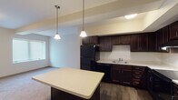 This single-floor unit is thoughtfully designed with a cohesive aesthetic carried throughout each space. The kitchen serves as a focal point, showcasing rich dark espresso cabinetry that extends to the ceiling, offering generous storage. Black appliances, including a refrigerator, range, and dishwasher, pair with light countertops to create a classic contrast. A central island with a matching countertop provides additional workspace and is illuminated by pendant lighting that adds warmth to the area. Wood-style flooring runs throughout the kitchen, transitioning to plush carpet in the adjacent living and bedroom areas.

The living space is open and bright, receiving natural light through windows fitted with blinds. The bedroom features soft carpeting, a tall narrow window, and a spacious walk-in closet equipped with wire shelving and hanging rods, providing ample organization options.

The bathroom is well-finished, featuring a dark vanity with drawers and cabinet storage, a white countertop, a lighted mirror, a medicine cabinet, and a walk-in shower with built-in shelving. Dark hardwood-style flooring completes the bathroom's polished look. The overall palette of warm neutrals and dark wood tones creates a consistent, inviting atmosphere throughout the unit.