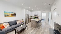 Located at 175 SW 7th Ave in Florida City, FL, this thoughtfully designed 2-bedroom, 3-bathroom unit offers a clean, modern aesthetic across a single floor. The open-concept living and kitchen area serves as the heart of the home, featuring light wood-look flooring, recessed lighting, and a seamless flow between spaces. The kitchen is outfitted with high-gloss white upper cabinetry, warm wood-toned lower cabinets, stone-look countertops, a decorative tile backsplash, and a full suite of stainless steel appliances. A central island with bar seating provides additional dining and prep space. The living area is bright and airy, with natural light filtering through slatted blinds. Both bedrooms are well-proportioned and finished with the same light flooring and neutral tones. The primary bedroom features sliding glass doors opening directly to a private outdoor area with a fenced yard and greenery. The second bedroom includes a large window with outdoor views. The three bathrooms are finished with modern tile work, including a walk-in glass-enclosed shower and a separate tub-shower combination. Additional features include closet storage, a dedicated hallway, and a secondary living or flex space with a window and television setup.