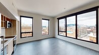 This contemporary urban residence is situated on an upper floor, offering sweeping city views from nearly every room. The layout is thoughtfully designed, beginning with a clean, well-lit hallway that connects the various living spaces. Light wood-tone luxury vinyl plank flooring runs throughout the main areas, complemented by plush carpeting in the bedrooms, creating a comfortable and cohesive aesthetic.

The living area is bright and open, featuring an expansive arrangement of large black-framed windows that frame impressive panoramic views of the surrounding cityscape, flooding the space with natural light. The crisp white walls and neutral tones throughout contribute to a clean, modern atmosphere.

The bedrooms are generously sized, each with soft carpeting underfoot and large windows overlooking the urban landscape. Closet space is integrated into the bedrooms, providing practical storage solutions.

The unit includes two bathrooms. The primary bathroom features a wide vanity with a white countertop, wood-toned cabinetry, a large mirror, and a glass-enclosed walk-in shower. The secondary bathroom offers a combined shower and tub, a single-sink vanity, and tile flooring. An in-unit stacked washer and dryer adds everyday convenience. Overall, the space delivers a polished, move-in-ready environment with a strong urban character.