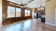 Located at 214 Broad St in Kingsport, Tennessee, this 2-bedroom, 2-bathroom residence showcases a compelling blend of industrial character and modern finishes. The open-concept living, dining, and kitchen area serves as the heart of the home, highlighted by exposed brick walls, polished hardwood flooring, and dramatic exposed ductwork overhead. A striking cast-iron column anchors the living space, while pendant chandeliers and ceiling fans add warmth and functionality. Large windows flood the area with natural light and offer views of the surrounding cityscape. The kitchen features dark cabinetry, granite countertops, and stainless steel appliances, including a refrigerator, range, microwave, and dishwasher. A hallway with louvered closet doors connects the private quarters, which include two well-proportioned bedrooms, each outfitted with ceiling fans and rich hardwood floors. The primary bedroom offers multiple closets for generous storage. Both bathrooms are finished with dark wood vanities, granite countertops, oval mirrors, and brushed nickel fixtures. Additional conveniences include a dedicated laundry room, utility room, and four closets throughout. The overall ambiance blends urban industrial aesthetics with comfortable, modern living.