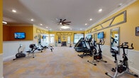 This single-level fitness center is designed to provide a comprehensive workout environment within an inviting, well-appointed space. The interior is immediately distinguished by its bold yellow walls accented with decorative gray geometric trim panels, white crown molding, and white wainscoting, creating a bright and energizing atmosphere. Recessed lighting and a large ceiling fan ensure the space remains well-lit and comfortable during use.

The main workout area is generously proportioned, accommodating a wide array of cardio equipment including treadmills, ellipticals, a stair climber, stationary bikes, and rowing machines. A dedicated strength training section features a cable crossover machine, a dumbbell rack with an assortment of free weights, and a workout bench. Wall-mounted flat-screen televisions and strategically placed mirrors enhance the exercise experience throughout the room.

Natural light filters in through multiple windows dressed with layered curtains, adding to the room's openness. The durable patterned carpet tiles covering the floor provide both comfort and practicality. An adjoining area, labeled "Play Room," includes a reception-style counter, additional equipment, and framed artwork, suggesting a multifunctional space suited for various activities. The overall ambiance is cheerful, clean, and purpose-built for active use.