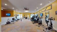 This single-level fitness center is designed to provide a comprehensive workout environment within an inviting, well-appointed space. The interior is immediately distinguished by its bold yellow walls accented with decorative gray geometric trim panels, white crown molding, and white wainscoting, creating a bright and energizing atmosphere. Recessed lighting and a large ceiling fan ensure the space remains well-lit and comfortable during use.

The main workout area is generously proportioned, accommodating a wide array of cardio equipment including treadmills, ellipticals, a stair climber, stationary bikes, and rowing machines. A dedicated strength training section features a cable crossover machine, a dumbbell rack with an assortment of free weights, and a workout bench. Wall-mounted flat-screen televisions and strategically placed mirrors enhance the exercise experience throughout the room.

Natural light filters in through multiple windows dressed with layered curtains, adding to the room's openness. The durable patterned carpet tiles covering the floor provide both comfort and practicality. An adjoining area, labeled "Play Room," includes a reception-style counter, additional equipment, and framed artwork, suggesting a multifunctional space suited for various activities. The overall ambiance is cheerful, clean, and purpose-built for active use.