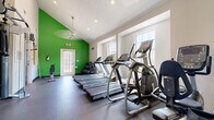 Located at 4211 Clay Hill Dr in Houston, Texas, this single-floor fitness center is designed to support a well-rounded workout experience. The space is defined by its striking vaulted ceilings, which lend an open, airy feel to the room, and bold green accent walls that energize the environment. Rubber flooring throughout provides both durability and comfort underfoot.

The cardio zone is well-equipped with a row of Matrix treadmills and elliptical trainers, positioned along a wall of windows that allow ample natural light to fill the space and offer views of the surrounding exterior. A recumbent stationary bike rounds out the cardiovascular offerings. A wall-mounted flat-screen television is installed near the center of the room for entertainment during workouts.

For strength training, the facility features a multi-station cable machine suitable for a wide range of resistance exercises, a dedicated free weight area with a full rack of dumbbells, and an adjustable weight bench. A ceiling fan keeps air circulating throughout the space, and recessed lighting supplements the natural light from the multiple windows. The fitness center is accessible via both a main interior entrance and a secondary exterior door, making it conveniently reachable from multiple access points.