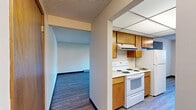 Located at 511 S Sullivan Rd in Spokane Valley, WA, this two-bedroom, two-bathroom residence offers a practical and well-organized single-floor layout. The living room is a generous, open space featuring gray vinyl plank flooring, a textured ceiling, baseboard heating, and a sliding glass door that leads to a private balcony overlooking the surrounding neighborhood and mature trees. The galley-style kitchen is efficiently designed with warm wood cabinetry lining both walls, white appliances including a range, refrigerator, and dishwasher, light vinyl plank flooring, a double basin sink, and a drop-panel ceiling with overhead lighting. A window at the end of the kitchen brings in natural light. Both bedrooms are carpeted and feature warm-toned wood closet doors, with the primary bedroom offering direct access to one of the two bathrooms. Each bathroom includes a wood-cabinet vanity, large mirror, and white countertop sink. One bathroom features additional overhead cabinetry for extra storage. A hallway with a dedicated closet connects the main living areas to the bedrooms. Throughout the unit, warm wood door frames provide visual continuity against neutral wall tones.